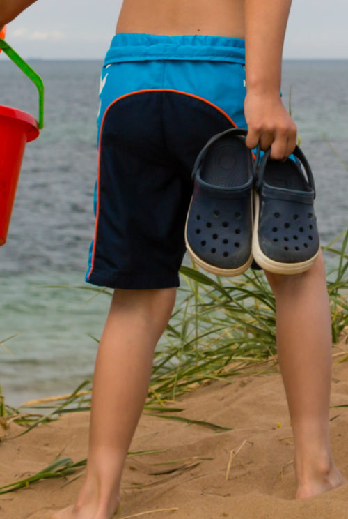 Cruise Packing List | Packing for a cruise - don't forget water shoes. Photo of boy walking with shoes in hand on shore.