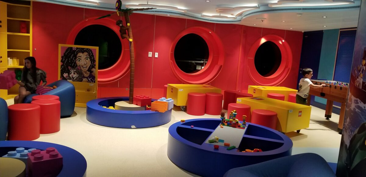 Circular blue LEGO building tables sit beneath red porthole-style wall openings and a decorative palm tree.