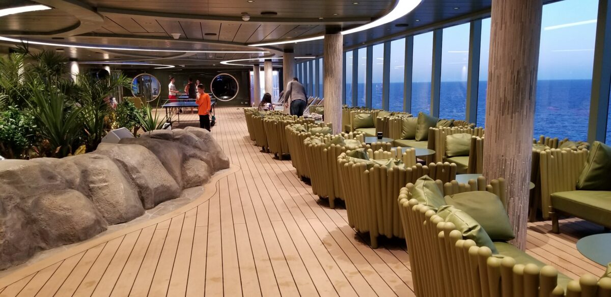 A wood-paneled deck with green tube-shaped lounge seating faces floor-to-ceiling windows overlooking the open ocean.