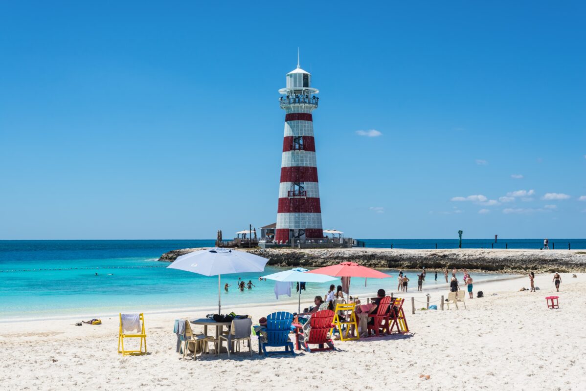 Beachgoers gather under umbrellas on white sand, with a rock jetty and lighthouse visible in the background.