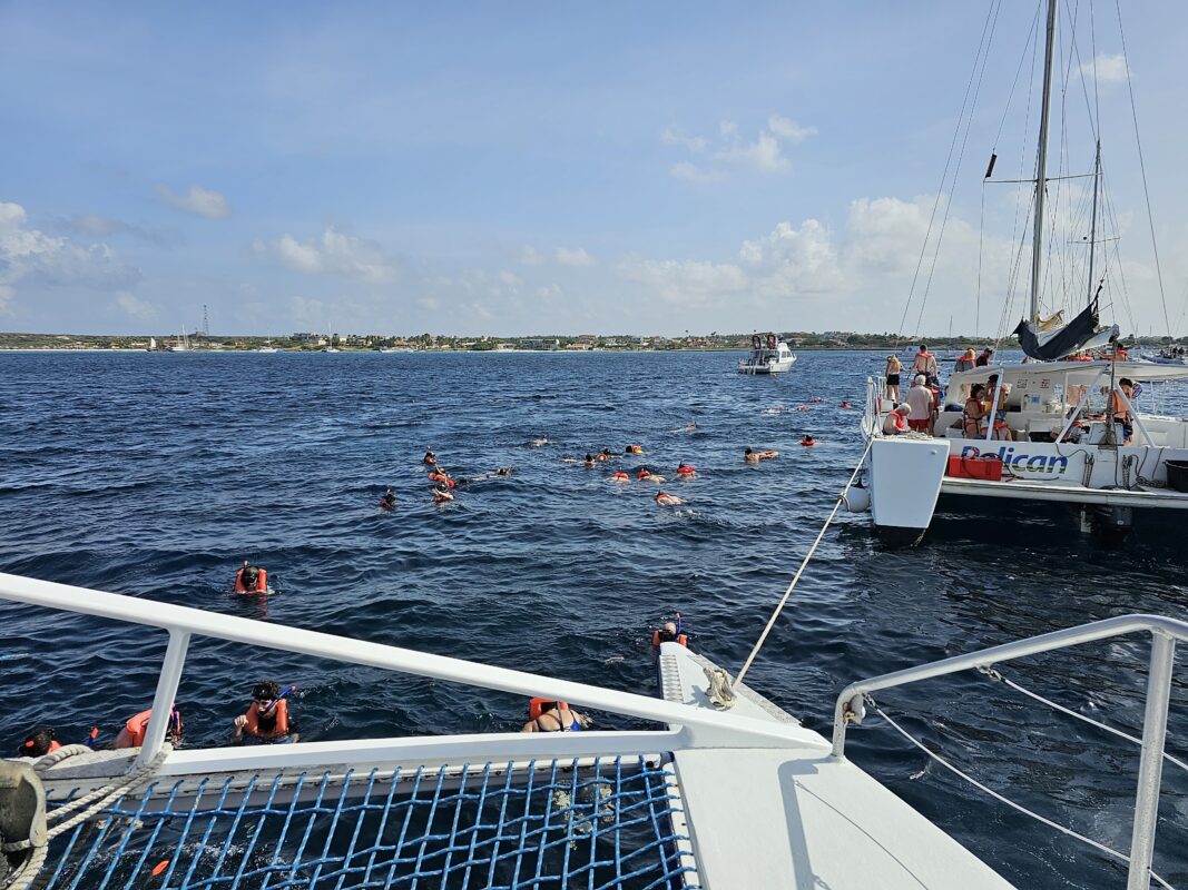 Snorkelers in life vests explore the waters near the Antilla shipwreck off Aruba, with catamarans anchored nearby and the shoreline in the distance.