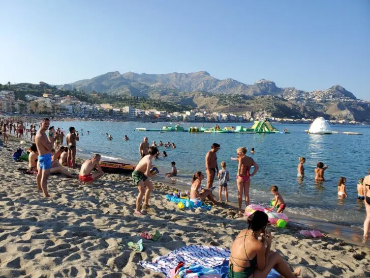 Crowded beach near Messina, Italy, with beachgoers enjoying the water and an inflatable water park, set against a backdrop of mountains and coastal buildings.