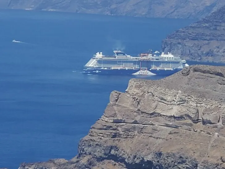 Celebrity Edge cruise ship anchored near the steep cliffs of Santorini, Greece, with the island's rugged, rocky landscape in the foreground and the deep blue waters of the caldera surrounding the ship.