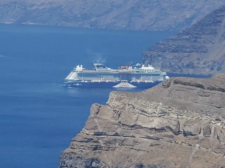 Celebrity Edge cruise ship anchored in the caldera of Santorini, Greece, with dramatic cliffs and blue waters in the background.