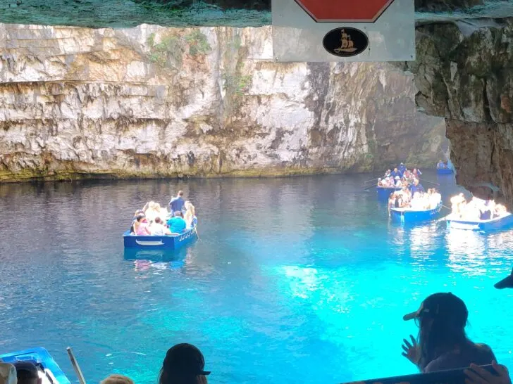 Tourists in rowboats explore the bright turquoise waters of Melissani Cave in Kefalonia, Greece.  Sunlight filters in through an opening above, illuminating the water below. Several boats filled with visitors float leisurely in the calm lake.