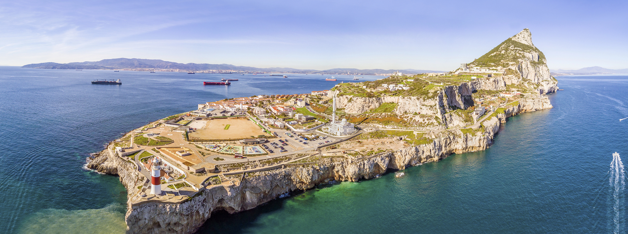 Aerial view of the Rock of Gibraltar with its limestone cliffs, lighthouse, and mosque, surrounded by the waters of the Mediterranean Sea, with ships passing through the Strait of Gibraltar.