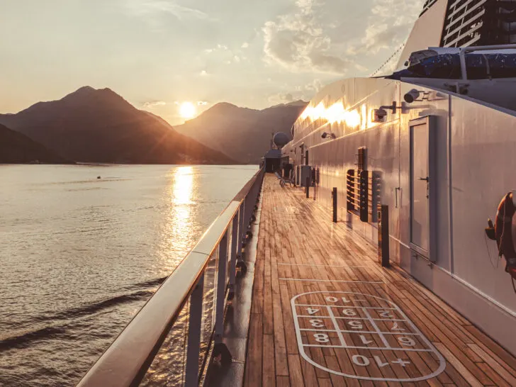 Best Mediterranean Cruise Lines | Cruise ship promenade deck at sunset with a shuffleboard court and mountain views in the background.