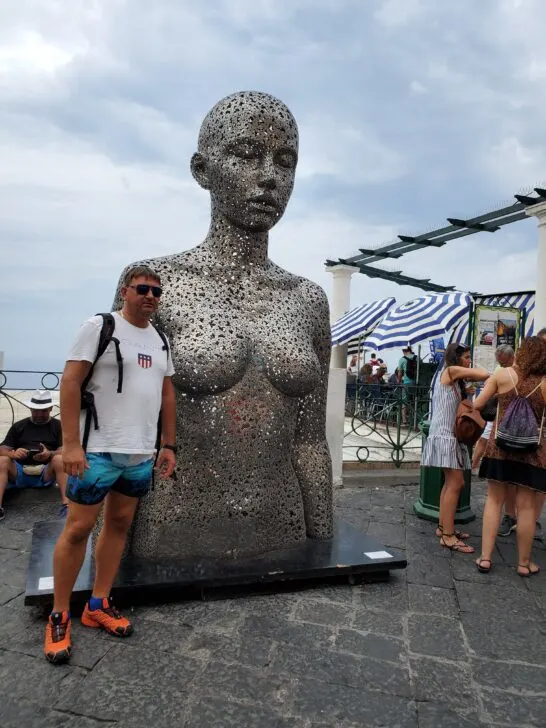 Metallic perforated sculpture of a human figure on display in Capri, Italy, with tourists in the background.