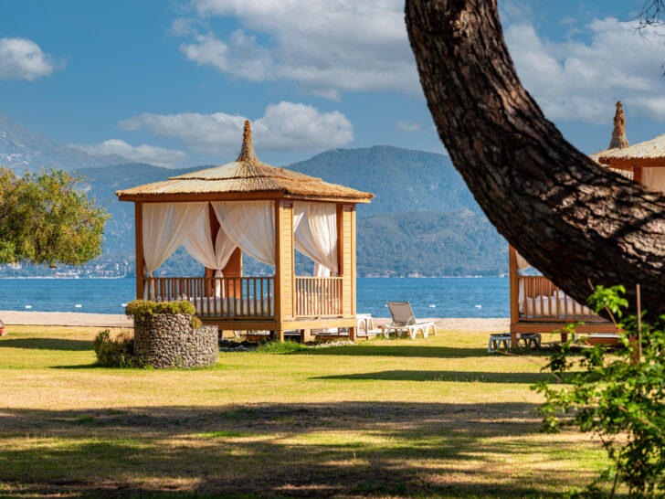 Mediterranean-style beach cabana with flowing curtains, set against a backdrop of mountains and the sea, located on a hotel property.