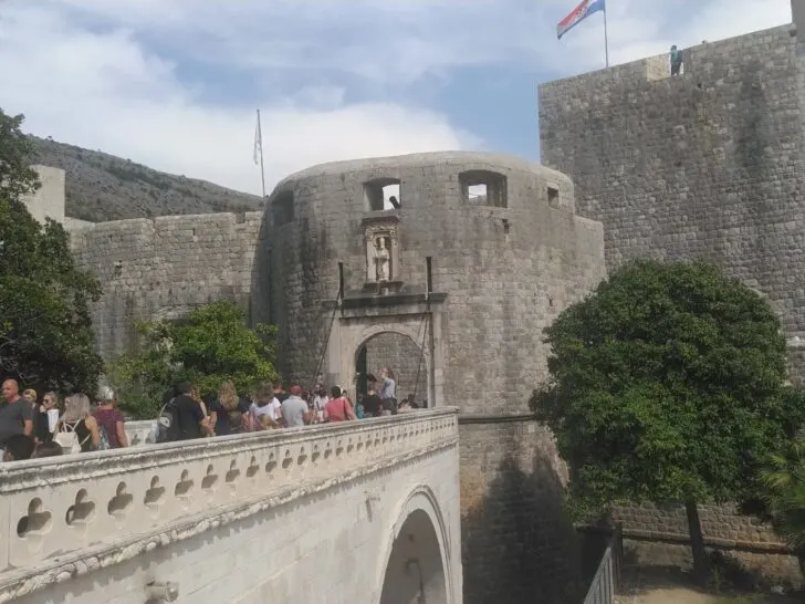 Tourists walking across a stone bridge leading to the ancient city walls of Dubrovnik, Croatia, with the Croatian flag flying above.