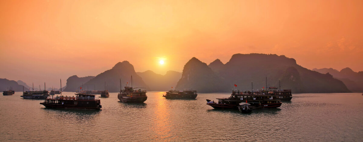 Traditional boats on the calm waters of Halong Bay, Vietnam, with a golden sunset reflecting off the sea and dramatic limestone karsts in the background, one of the world’s most iconic and unique cruise destinations.