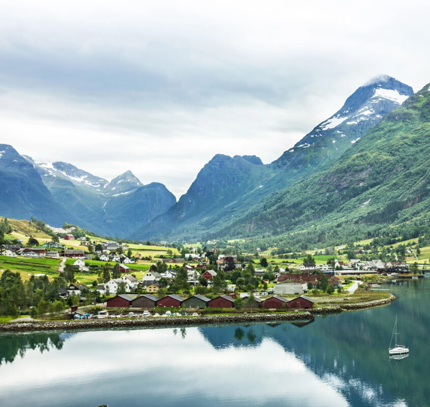 A scenic view of a Norwegian village along a calm fjord, surrounded by towering green mountains and snow-capped peaks, showcasing one of the most unique cruise destinations in Europe.