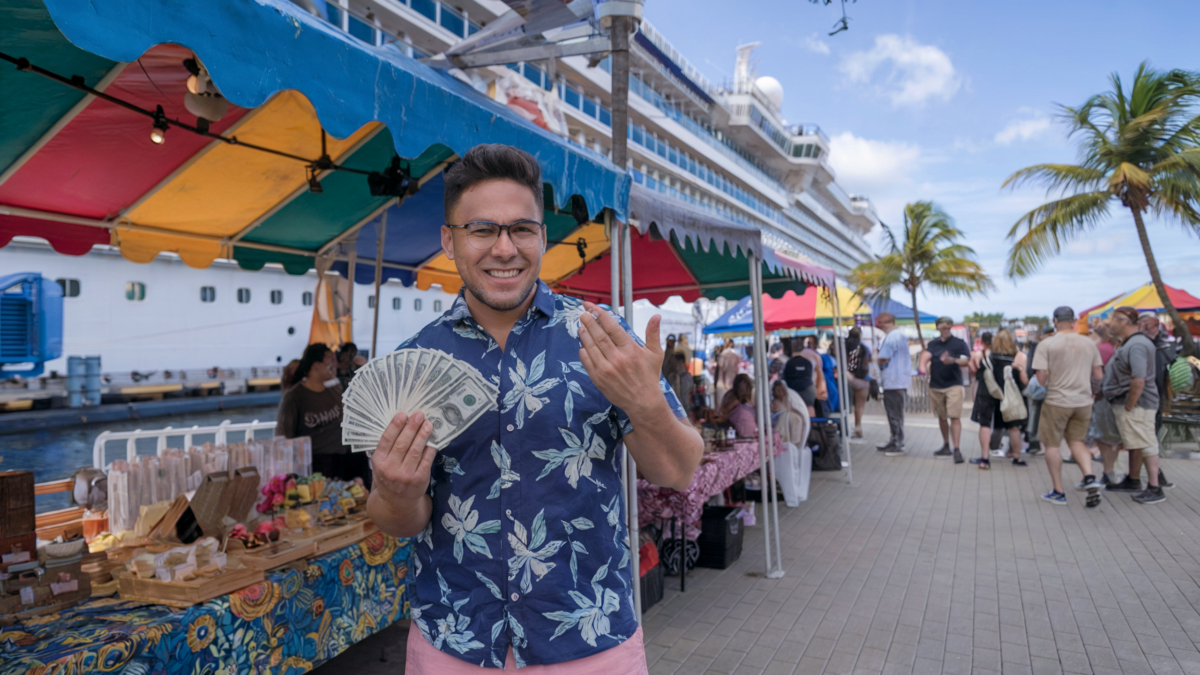 A smiling man in a colorful marketplace near a cruise ship holding a fan of cash, illustrating the potential for cruise scams during excursions.