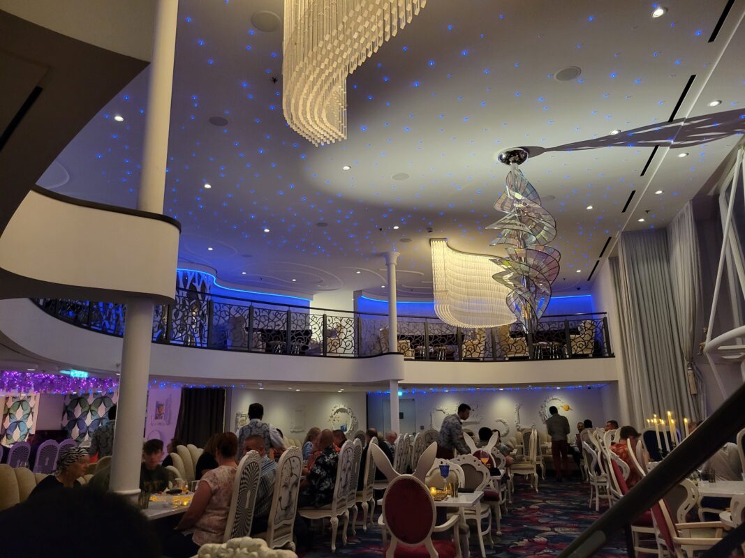 A whimsical two-story dining room at Wonderland, a specialty restaurant on Royal Caribbean’s Symphony of the Seas. The ceiling is illuminated with glowing blue lights, and the decor features white and gold chandeliers, abstract sculptures, and ornate chairs inspired by the Alice in Wonderland theme.