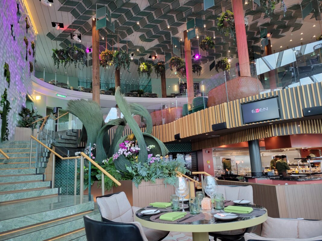 A vibrant, nature-inspired dining space at Eden restaurant on Celebrity Edge-class ships. The multi-level venue features hanging greenery, curved architectural elements, and an open kitchen where chefs prepare specialty dishes.