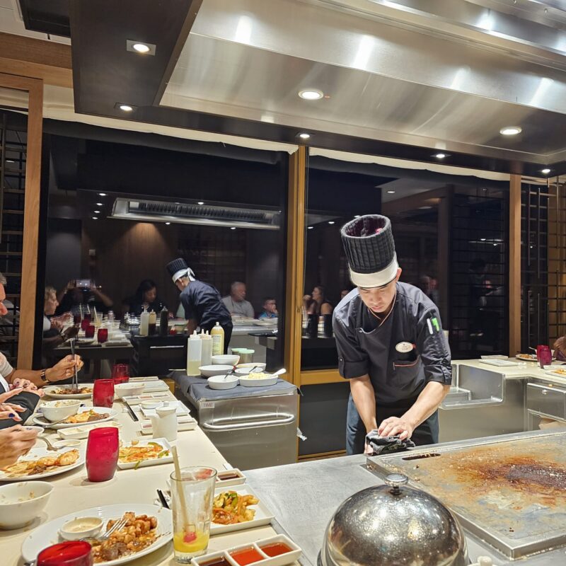 A chef at Izumi Teppanyaki on Wonder of the Seas prepares hibachi-style cuisine for cruise guests in a lively specialty dining setting.