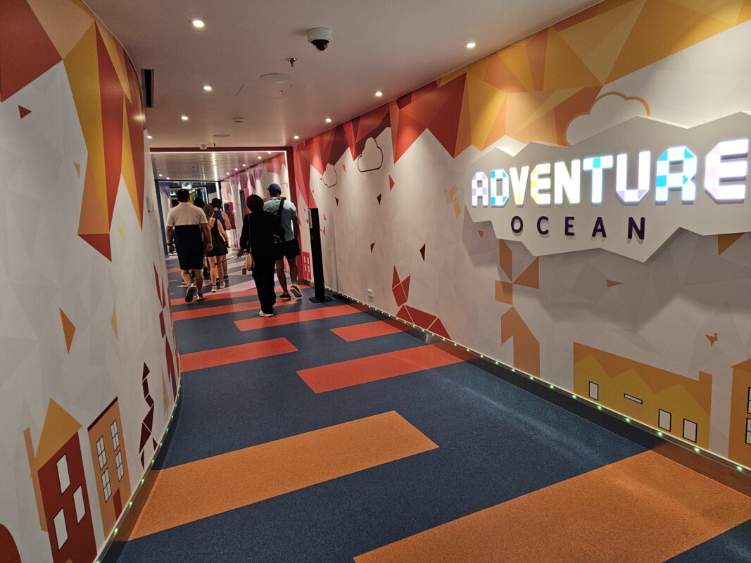 Vibrant corridor with geometric designs and an illuminated ‘Adventure Ocean’ sign, marking the entrance to a children’s play area on a cruise ship.