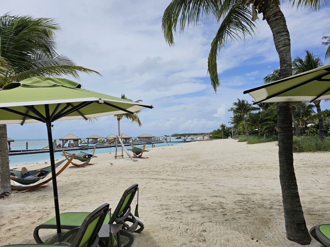 A view of Coco Beach Club at CocoCay, featuring green umbrellas, lounge chairs, hammocks, and private overwater cabanas along the turquoise shoreline.