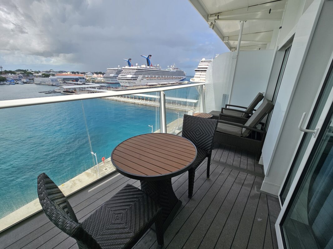 View from a cruise ship balcony showing a serene port with calm blue waters, docked ships, and a seating area with a table and lounge chairs.