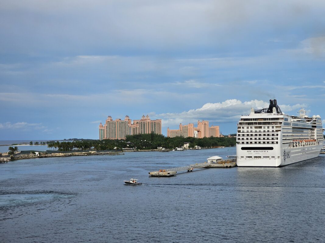 A large cruise ship, MSC Magnifica, is docked at the port in Nassau, Bahamas. In the background, the pink towers of Atlantis Paradise Island Resort rise above lush greenery, with calm blue waters and small boats visible in the harbor.