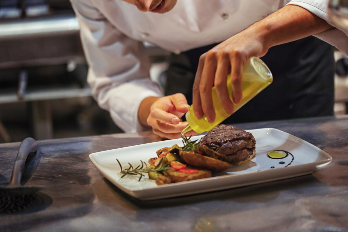 Best Specialty Dining | A chef in a white uniform carefully drizzles oil onto a plated gourmet steak dish, garnished with fresh herbs, roasted vegetables, and decorative sauce accents. 