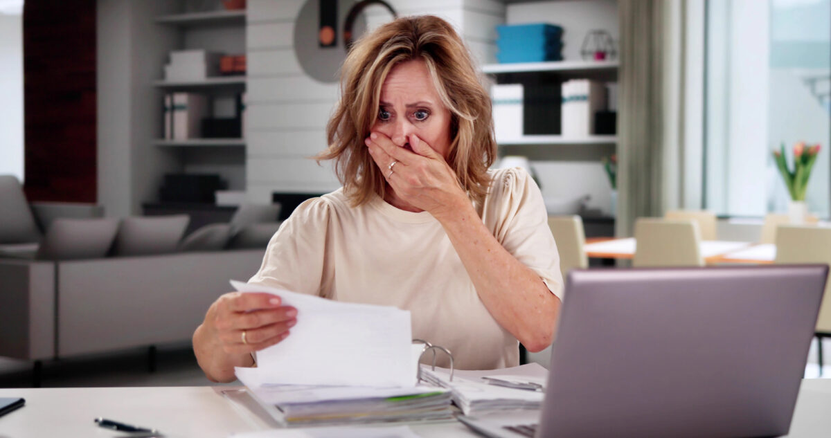 A shocked woman sitting at a desk, covering her mouth with her hand while looking at a bill.