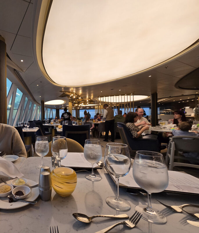 A specialty restaurant on a cruise ship featuring elegant décor, panoramic windows, and families enjoying dinner.