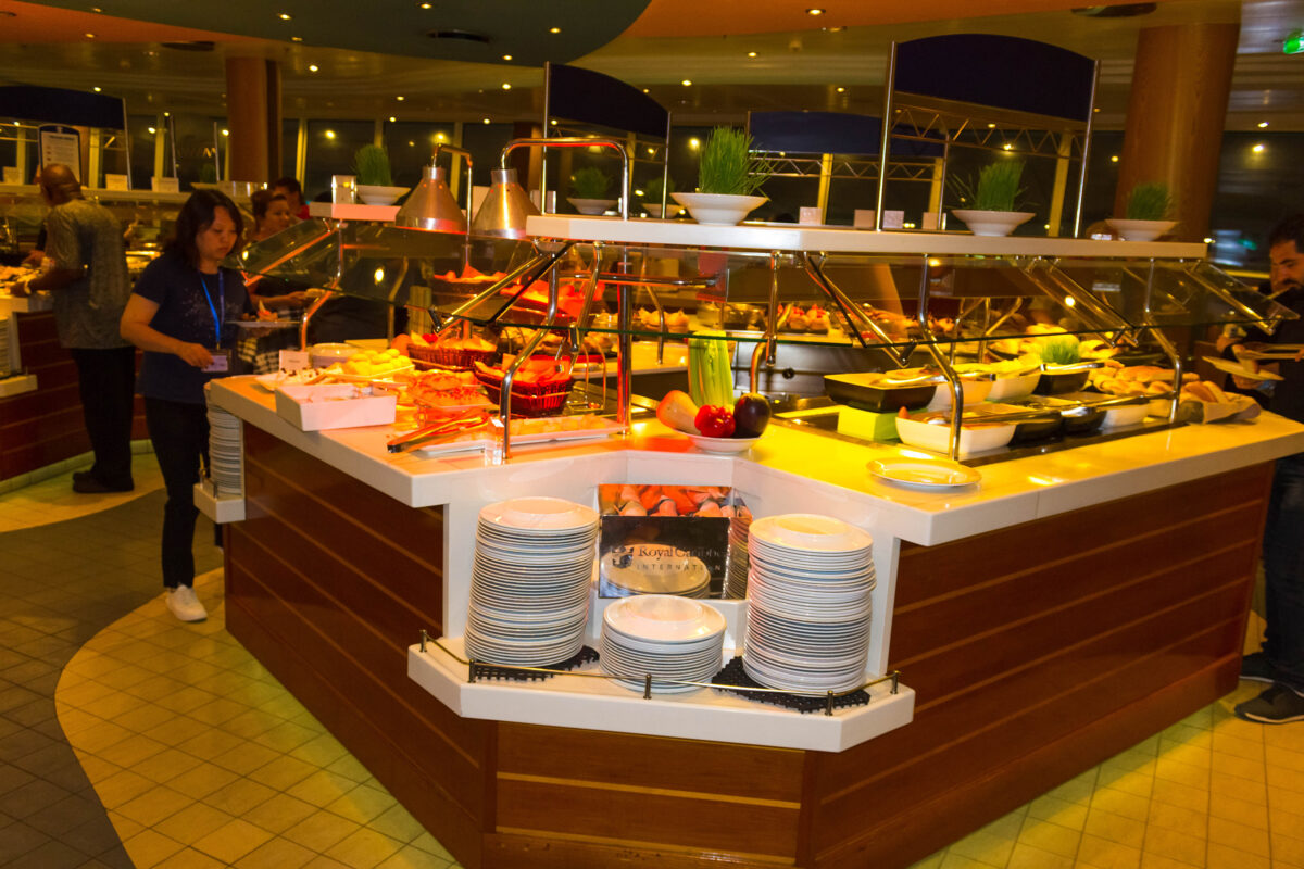 Cruise ship buffet station with a variety of food options and plates stacked in the foreground, with guests selecting their meals.