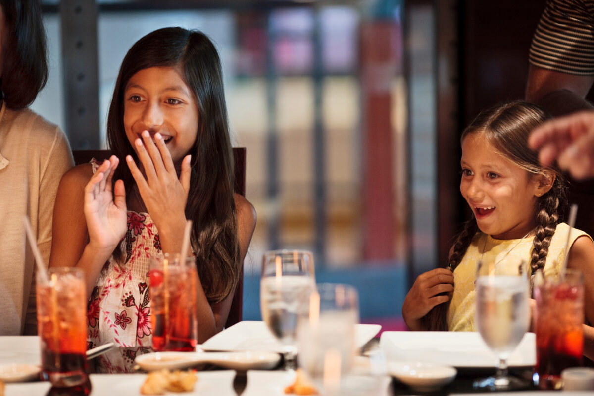 Two young girls smiling and excited while dining at a restaurant, with drinks and plates on the table.