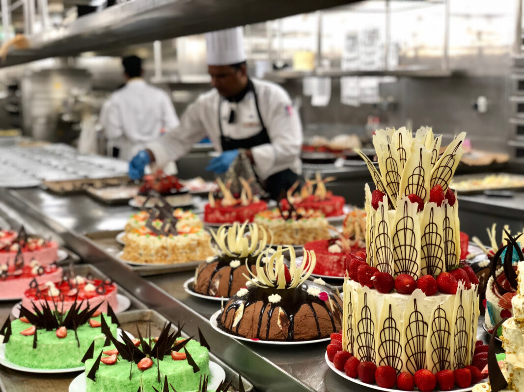 An array of intricately decorated cakes and pastries in a commercial kitchen, with a chef preparing fresh desserts in the background.