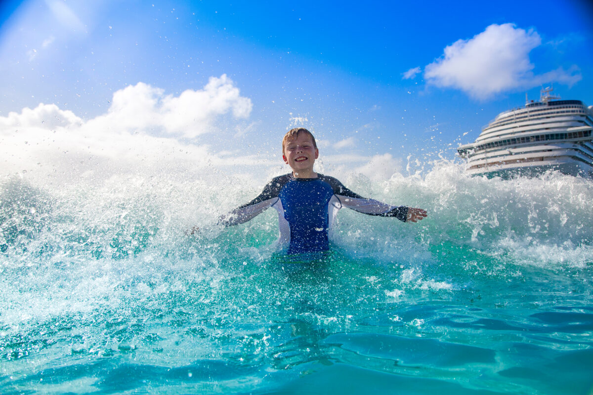 Spring Break Cruise Destinations | A young boy wearing a blue rash guard smiles as he splashes in the turquoise waters of the Caribbean with a cruise ship in the background under a bright blue sky.