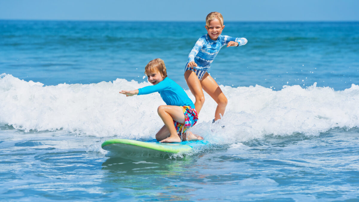 A young boy and girl in rash guards laugh as they balance on a surfboard, learning to surf in shallow ocean waves.