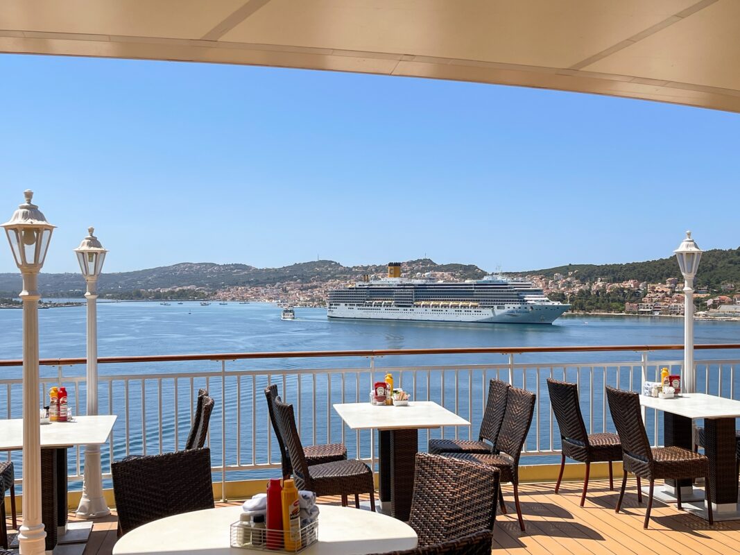 Outdoor dining area on a cruise ship with a view of the ocean, a distant cruise ship, and a coastal town.