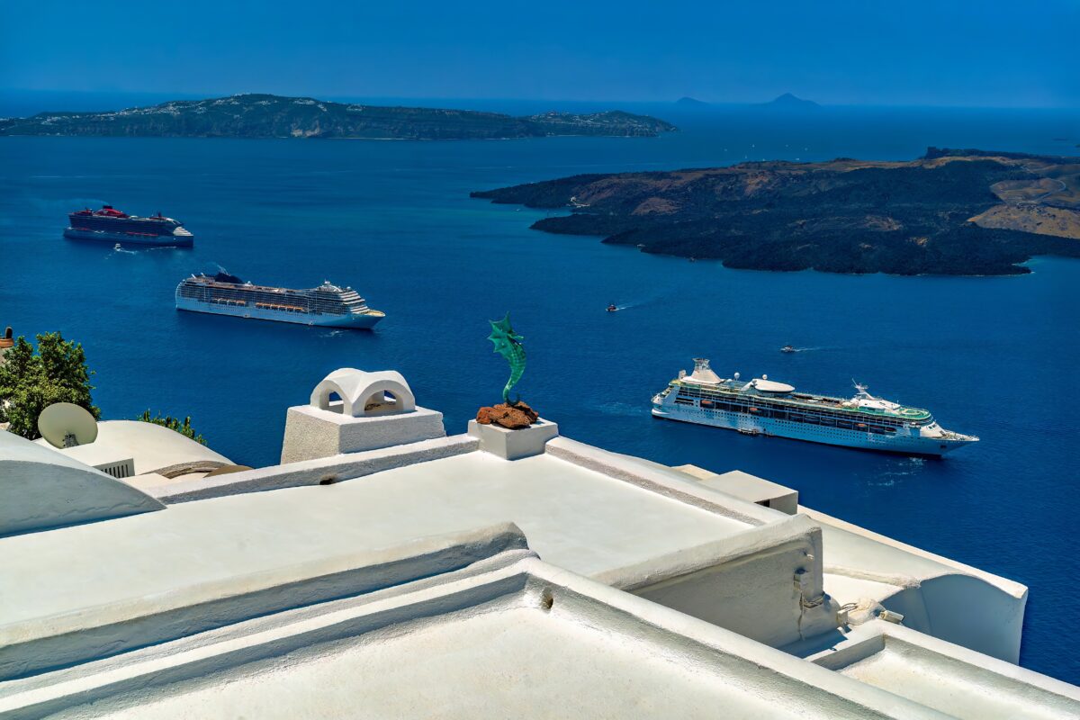 Cruise ships floating in the calm Mediterranean waters surrounded by rugged islands, as seen from a rooftop featuring white stucco and classic Greek details.