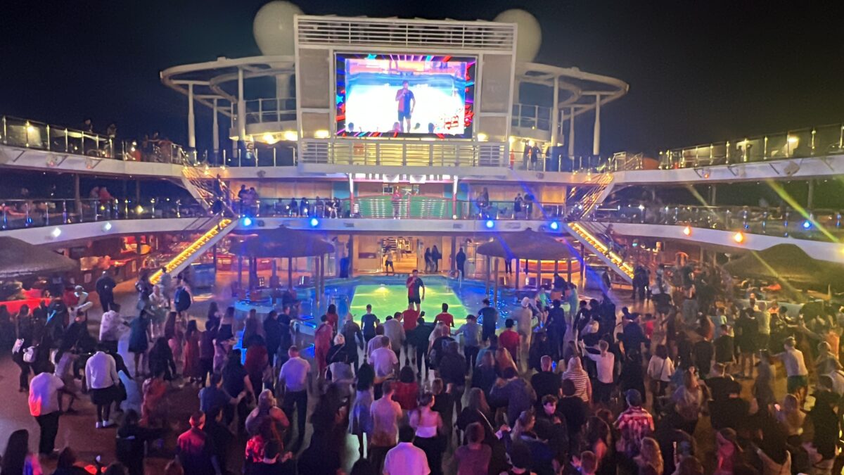 A lively nighttime party on a cruise ship’s pool deck, with a crowd dancing and a large outdoor screen displaying entertainment.