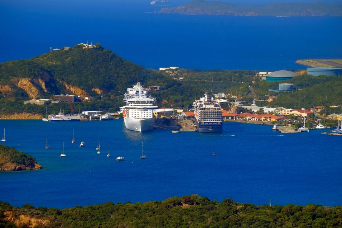 Caribbean Cruise vs Beach Resort | A Caribbean cruise port with two large cruise ships docked, surrounded by blue waters, green hills, and anchored sailboats, showcasing the appeal of cruise travel.