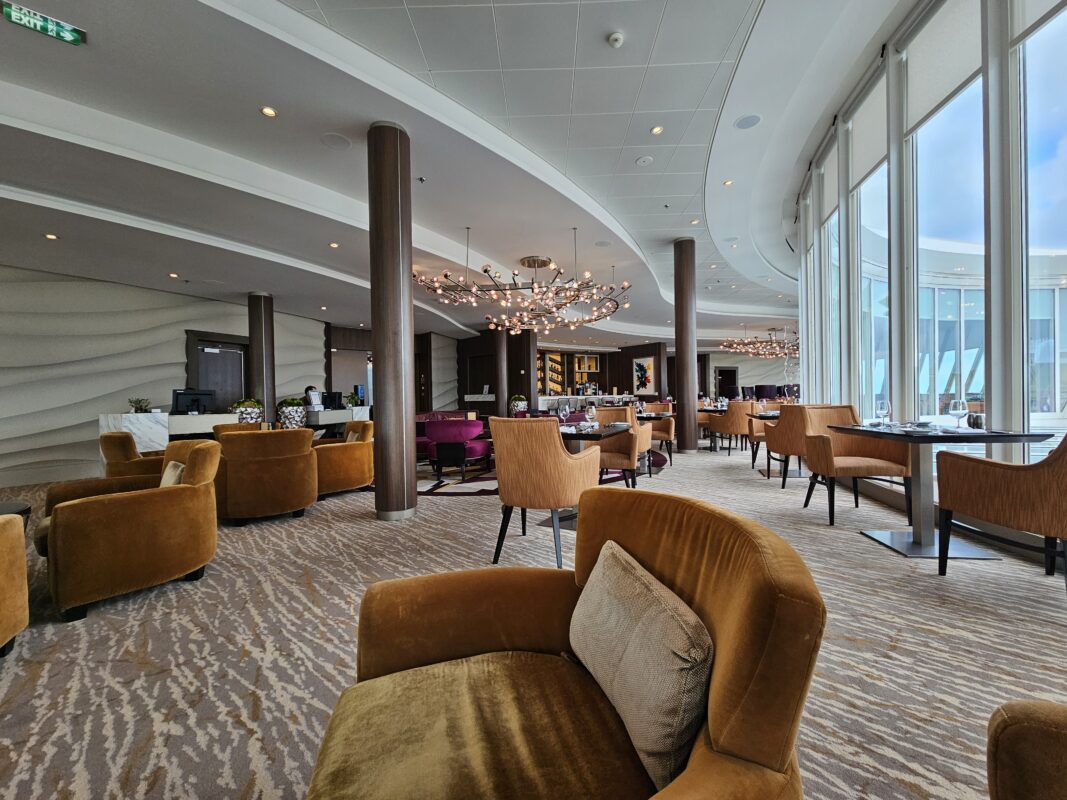 A spacious and elegantly designed private suite lounge on a Royal Caribbean ship, featuring plush mustard-colored armchairs, modern chandeliers, and a contemporary bar area with dining tables in the background.