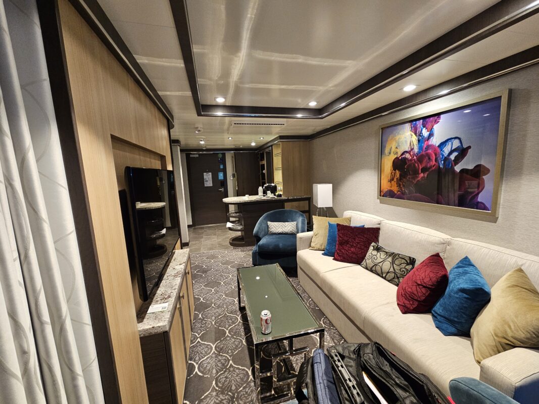 The separate living room of a grand suite on Symphony of the Seas including large couch, coffee table, media center, side chairs and bar area.