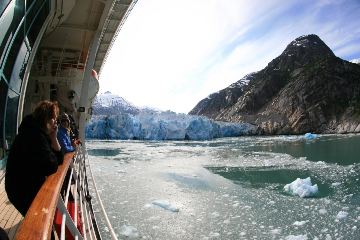 Cruise ship passengers stand at the railing, admiring a massive blue glacier surrounded by floating ice in Alaska.