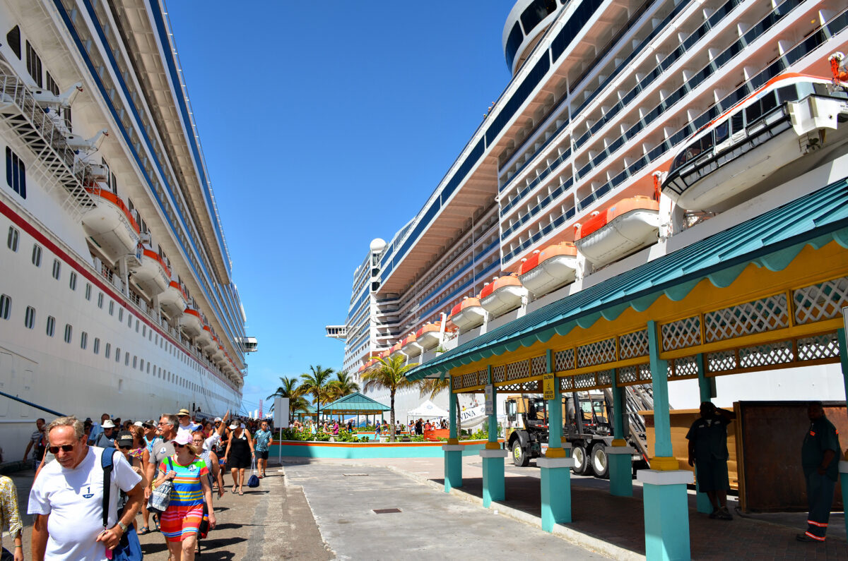 Cruise passengers walking along the pier in Nassau, Bahamas, between two large docked ships.