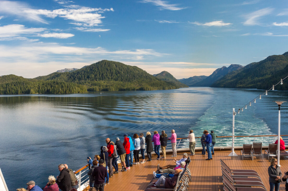 Alaska Cruise Worth It | A group of travelers stands along the railing of a cruise ship, admiring the breathtaking Alaskan scenery as the ship sails through a narrow waterway.