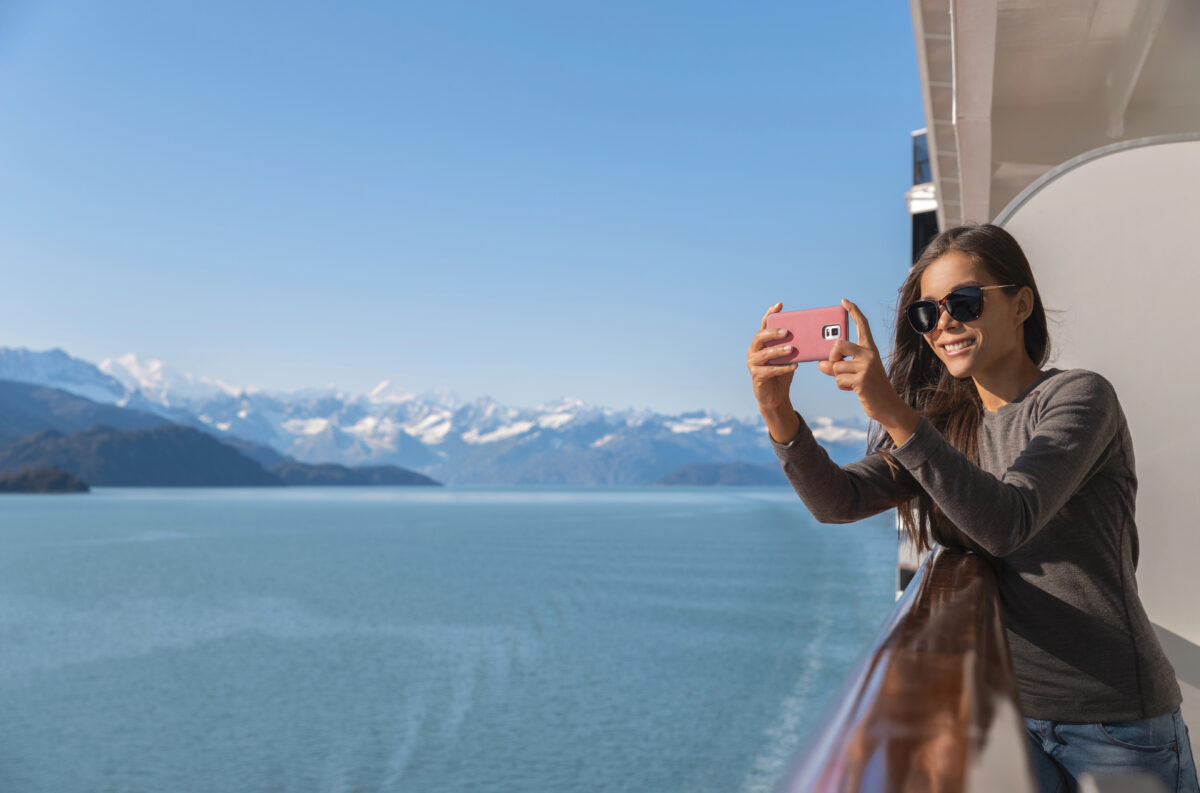 A cruise passenger leans over the railing, capturing Alaska’s breathtaking landscapes—one of the top reasons some think that a balcony is the best cabin for an Alaska cruise.