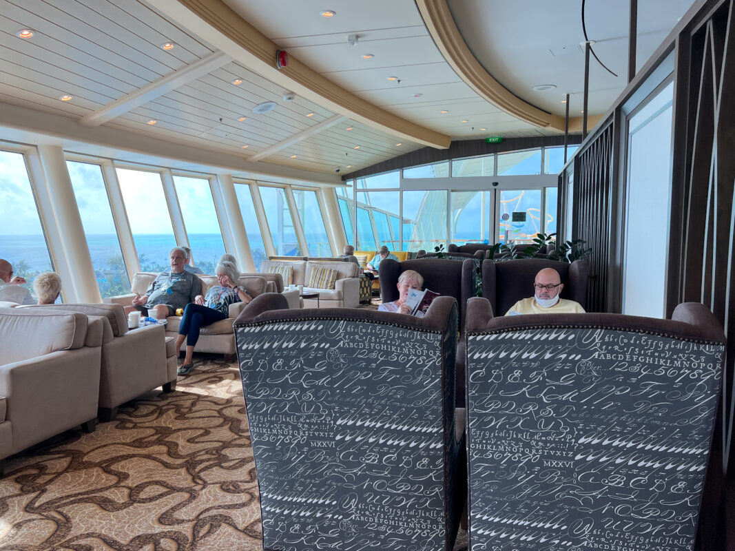 Spacious cruise ship lounge with panoramic ocean views. Passengers relax in plush seating, some enjoying drinks and reading, while natural light fills the space through large windows.