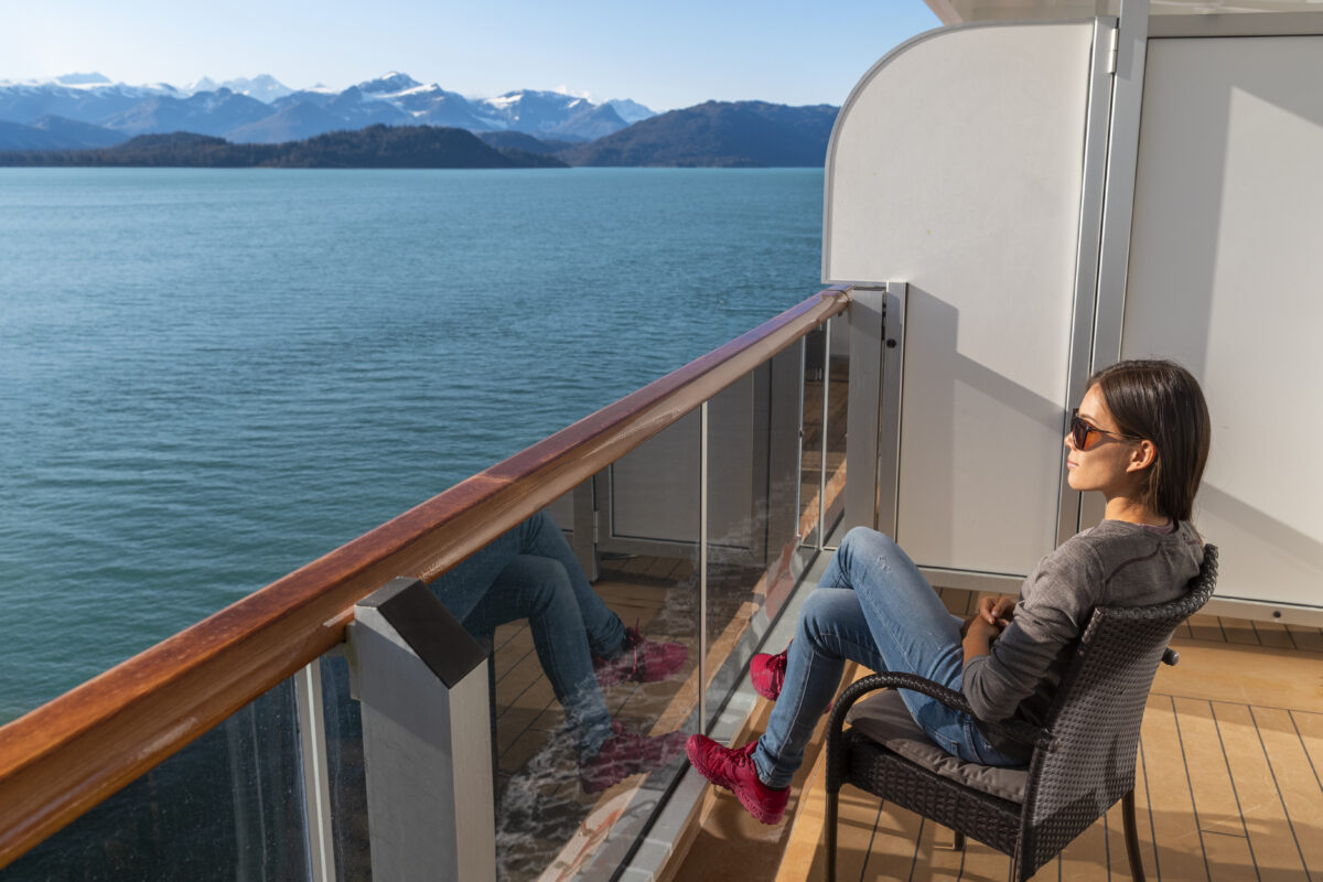 A woman sits on a cruise ship balcony, enjoying panoramic mountain and ocean views, showcasing why a balcony cabin is the best cabin for an Alaska cruise.