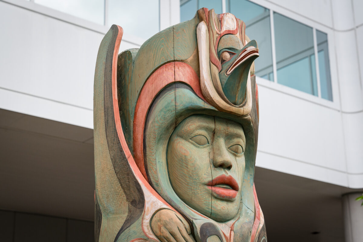 A close-up of a traditional Tlingit totem pole in Alaska, featuring intricate carvings of a human face and a bird figure.