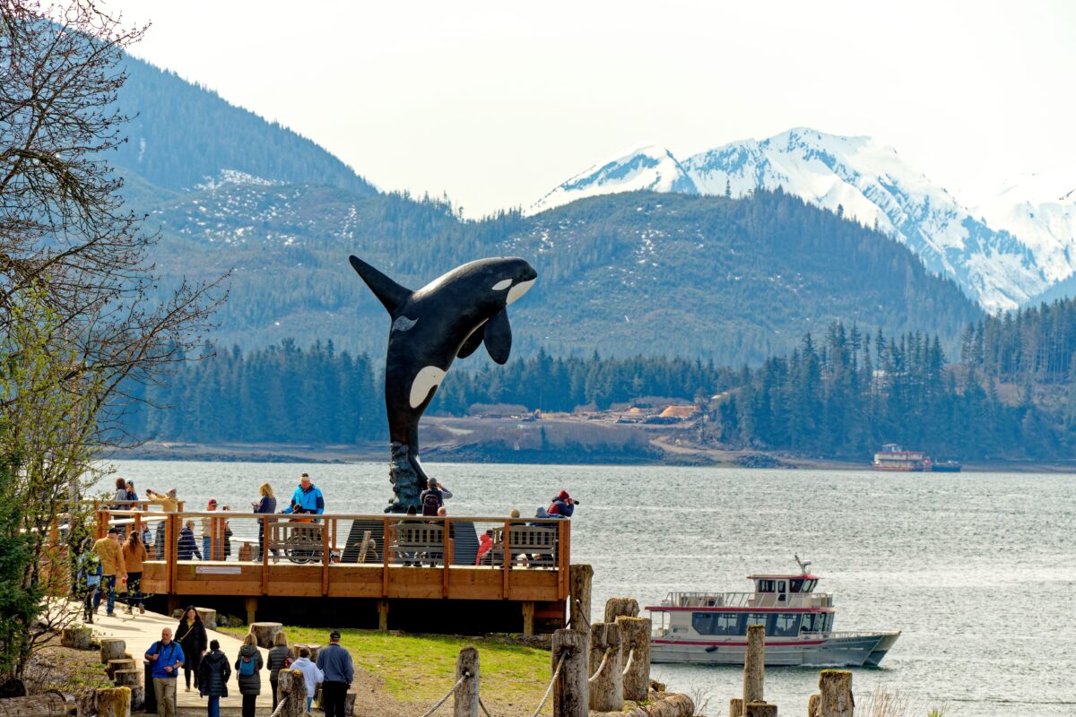 Tourists walking along a wooden boardwalk at Icy Strait Point, Alaska, with a large breaching orca statue in the foreground and snow-capped mountains in the background.