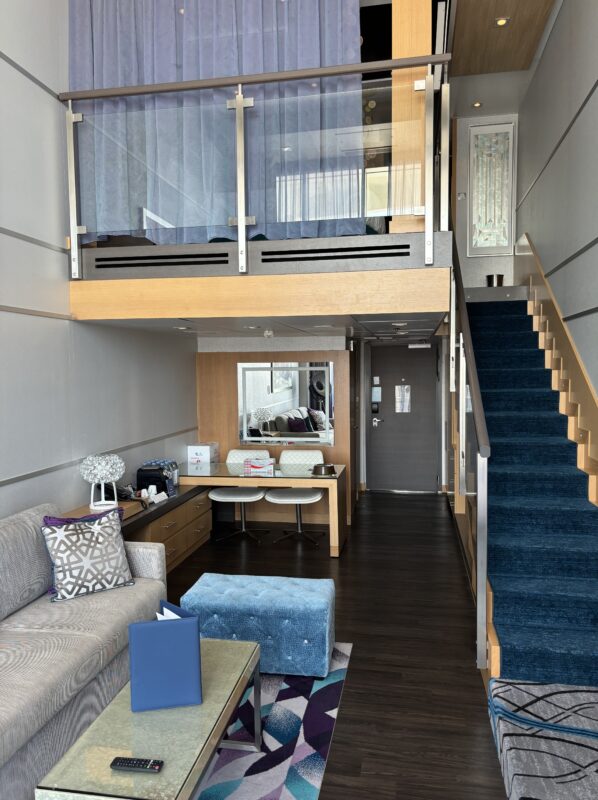 Spacious two-story cruise suite with modern furnishings. A luxurious loft-style stateroom features a cozy living area, a staircase leading to an upstairs bedroom, and sleek wood and glass accents.