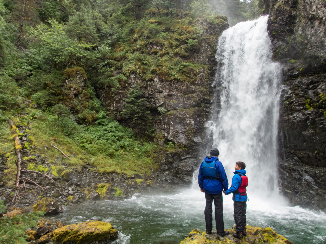 A father and son in blue rain jackets stand on a mossy rock, admiring a powerful waterfall in the Alaskan wilderness.