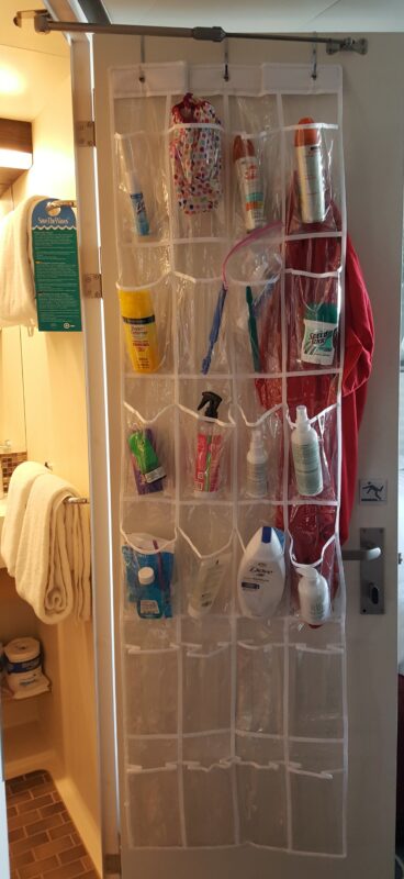 Clear plastic over-the-door shoe organizer hanging on a cruise cabin bathroom door, with multiple pockets holding various toiletry items including sunscreen, deodorant, toothbrushes, and body wash. This photo demonstrates a popular cruise cabin hack that maximizes vertical storage and keeps small bathrooms tidy and accessible for families.