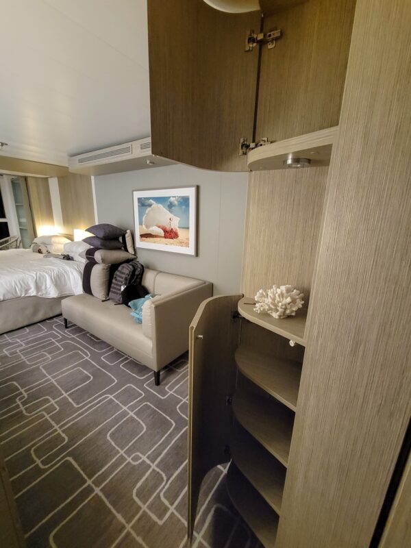 Cruise cabin interior with a modern beige-and-gray color scheme. In the foreground, a curved vertical cabinet is open to reveal several hidden corner shelves. In the background, a bed with stacked pillows, a loveseat with a towel animal, and a beach-themed wall photo are visible. This image shows an example of concealed storage often found in cruise cabins.